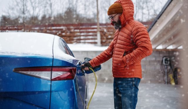 Man charging electric car during cold snowy day. Side view of hansome man putting charger in charging port during winter, cold weather. Charging and driving electric vehicles during winter season.